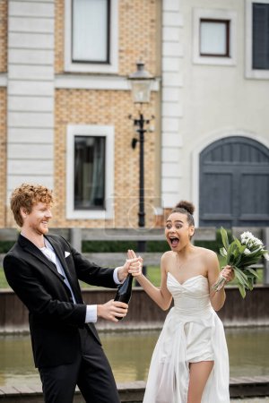 Hochzeit in der Stadt, rothaariger Bräutigam mit Champagner, der in der Nähe der schreienden afrikanisch-amerikanischen Braut aus der Flasche strömt
