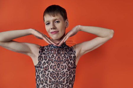 cheerful androgynous person smiling and posing on orange backdrop, hands near face, queer fashion