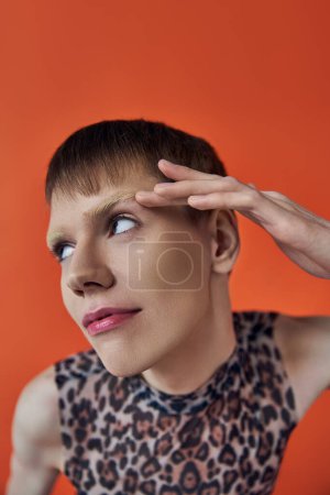 nonbinary person smiling and looking away on orange backdrop, touching eyebrow, queer fashion