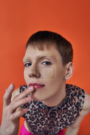 nonbinary person smiling and touching lips on orange backdrop, looking at camera, queer fashion