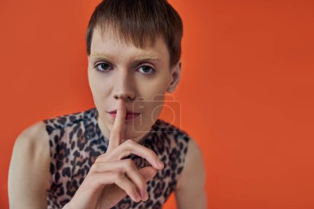 androgynous person in leopard print outfit showing hush sign on orange backdrop, queer fashion