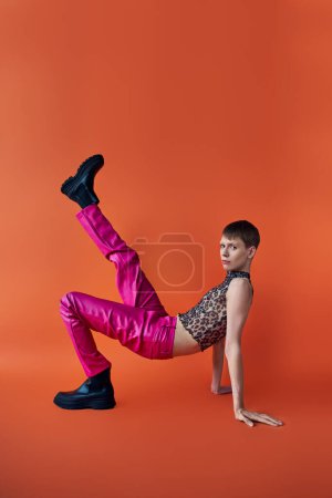 queer person in leopard print sleeveless top and pink pants posing on orange backdrop, fashion