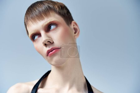 portrait, modèle androgyne avec maquillage regardant loin sur fond gris, beauté et visage, queer