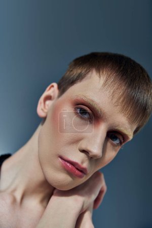 Queer Model mit Make-up posiert auf grauem Hintergrund, Schönheit, androgyn, Blick in die Kamera, Nahaufnahme