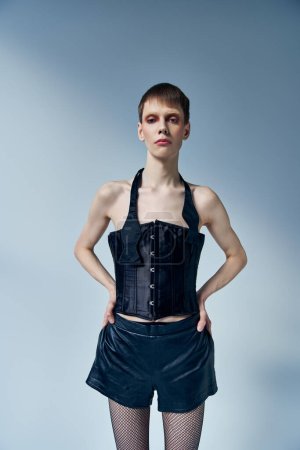queer model in black corset and shorts posing with hands on hips on grey backdrop, androgynous