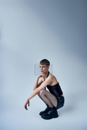 Queer Model in schwarzem Korsett und Netzstrumpfhose auf grauem Hintergrund sitzend, androgyne Person, Mode
