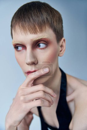 modelo queer con maquillaje mirando hacia otro lado sobre fondo gris, persona andrógina, retrato, identidad