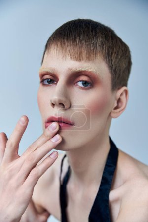 Photo for Queer person with makeup on grey backdrop, androgynous person, touching lip, look at camera - Royalty Free Image