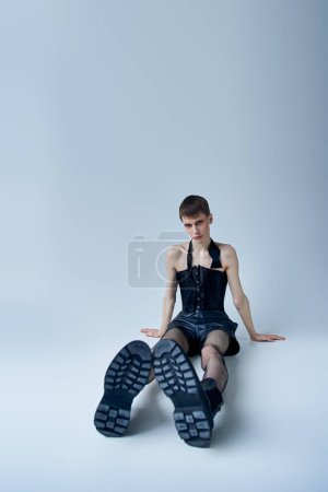 Téléchargez les photos : Modèle queer en corset noir et collants résille assis sur gris, personne androgyne, mode lgbt - en image libre de droit