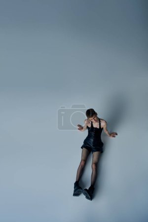 Androgynes Modell in Korsett und Netzstrumpfhose, sitzend auf grauem Hintergrund, queerer Stil, hoher Winkel