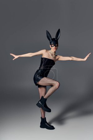 androgynous model in corset posing in bdsm bunny mask on grey, queer fashion, provocative outfit
