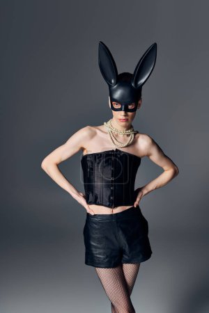 fashion, genderqueer person in corset posing in bdsm bunny mask on grey, queer style, hands on hips