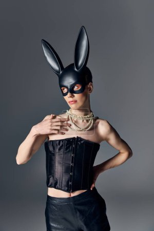 Mode, genderqueer Person im Korsett posiert in bdsm Hasenmaske auf grau, queer Stil, Hand auf Hüfte