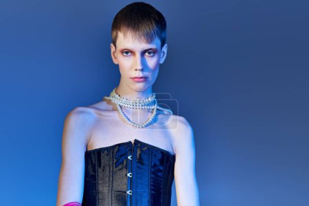 queer person in black corset and pearl necklace posing on blue backdrop, edgy fashion, nonbinary