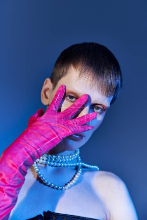 modelo queer en collar de perlas que cubre la cara con la mano en guante rosa sobre fondo azul, estilo vanguardista
