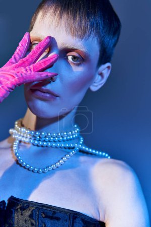 Foto de Modelo queer en collar de perlas que cubre la cara con la mano en guante rosa sobre fondo azul, moda audaz - Imagen libre de derechos