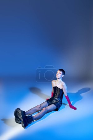 modelo queer en corsé y botas sentado sobre fondo azul, persona no binaria en guantes de color rosa, la moda
