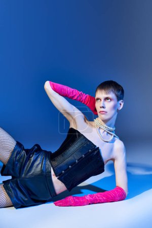 queer person in bold outfit and pink gloves posing on blue backdrop, shorts, nonbinary model, style