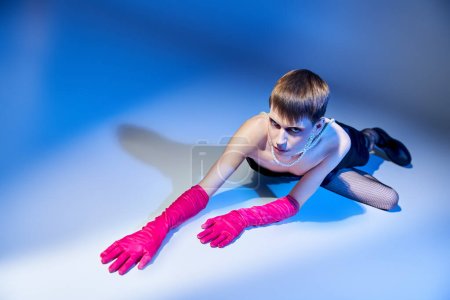 queer model in bold outfit and pink gloves posing on blue backdrop, nonbinary and extravagant