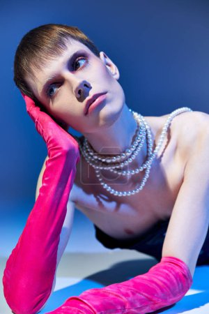 dreamy queer model in bold outfit and pink gloves posing on blue backdrop, nonbinary, look away