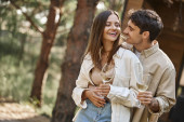 Smiling brunette man holding glass of wine and hugging girlfriend near vacation house outdoors Tank Top #669760872