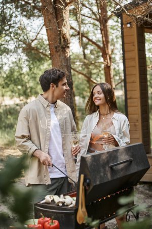Brunette homme cuisine sur le gril et parler à la petite amie souriante avec du vin près de la maison de vacances