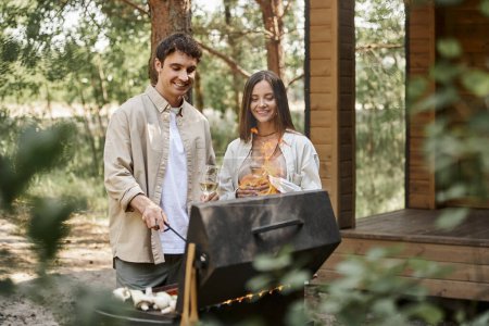 Smiling couple with wine cooking on grill while resting near vacation house at background