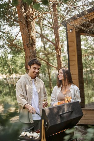 Fröhliches Paar mit Wein am Grill beim Picknick in der Nähe des Sommerhauses im Hintergrund