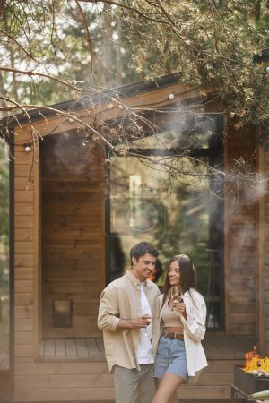 Alegre y elegante pareja sosteniendo el vino y de pie cerca de la parrilla con humo y casa de vacaciones
