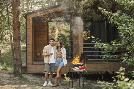 Positiva pareja romántica sosteniendo el vino y de pie cerca de la parrilla y la casa de vacaciones en el fondo
