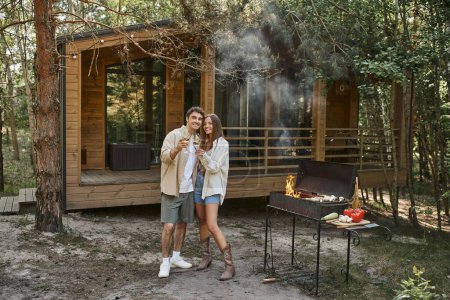 Téléchargez les photos : Couple romantique positif tenant du vin et regardant la caméra près du gril et de la maison de vacances - en image libre de droit