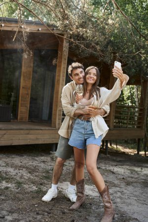 Téléchargez les photos : Homme souriant étreignant petite amie avec du vin prendre selfie sur smartphone près de la maison de vacances en plein air - en image libre de droit