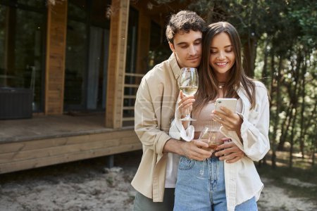 Stylish man hugging girlfriend with wine using smartphone near vacation house at background