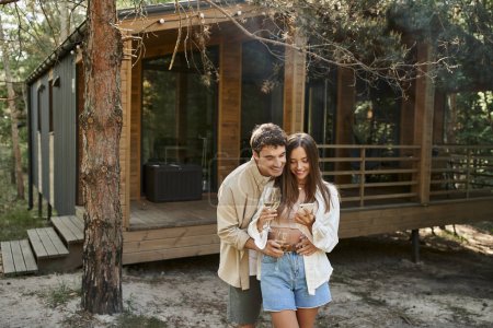 Téléchargez les photos : Couple romantique avec vin souriant tout en utilisant smartphone près de la maison de vacances en plein air - en image libre de droit
