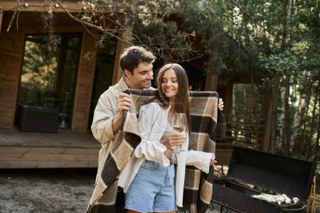 Man holding blanket near girlfriend with wine, grill and vacation house at background