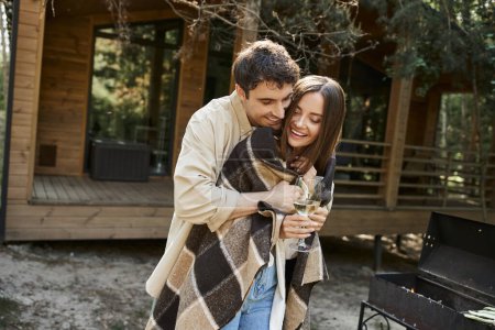 Cheerful man hugging girlfriend in blanket with wine near grill and vacation house at background