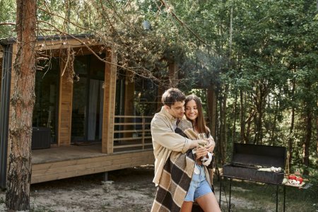 Man hugging smiling girlfriend with blanket and wine while standing near grill and vacation house