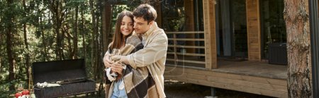Positive man hugging girlfriend in blanket with wine near grill and summer house, banner