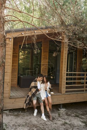 Téléchargez les photos : Couple romantique positif dans la couverture tenant du vin près du fromage tout en étant assis sur le porche de la maison d'été - en image libre de droit