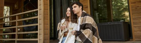 Téléchargez les photos : Couple souriant dans la couverture tenant le vin et regardant loin près de la maison de vacances à l'extérieur, bannière - en image libre de droit