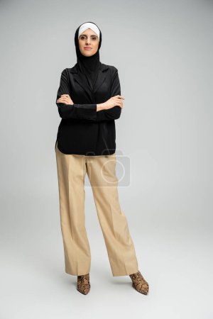stylische muslimische Geschäftsfrau in Blazer, Hose und Hijab posiert mit verschränkten Armen auf grau, durchgestreift