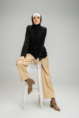 Geschäftsfrau in muslimischem Hijab und trendiger Freizeitkleidung sitzt auf einem hohen Hocker auf grauem, durchgestreiftem Boden