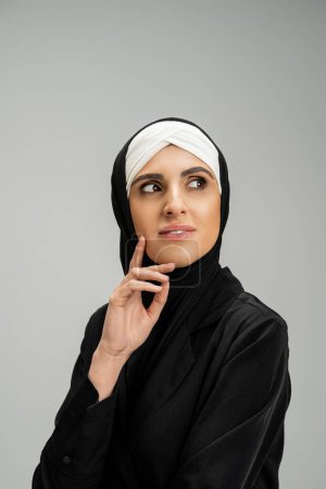 femme d'affaires musulmane souriante en hijab et veste noire regardant loin avec la main près du visage sur gris