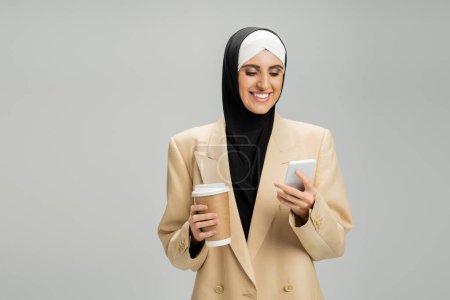 empresaria musulmana motivada en hijab y chaqueta, con teléfono inteligente y café para ir en gris
