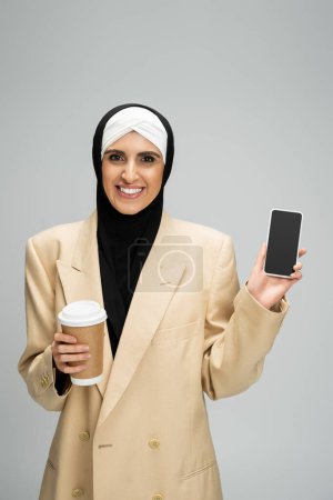 lächelnde, trendige muslimische Geschäftsfrau mit Coffee to go zeigt Smartphone mit leerem Bildschirm auf grau