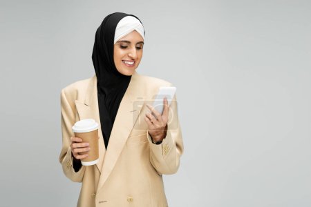 glücklich und trendy muslimische Geschäftsfrau mit Imbissgetränk Vernetzung auf Smartphone auf grau