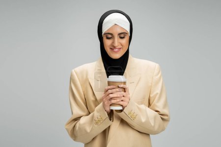 complacido mujer de Oriente Medio en hijab musulmán y chaqueta beige sosteniendo café para ir en gris