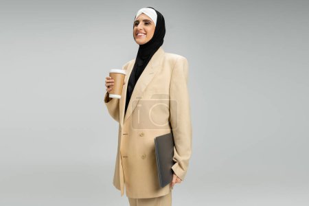 progresista y elegante mujer de negocios musulmana con bebida para llevar y portátil sonriendo en gris