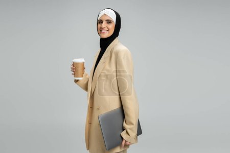 moderna y elegante mujer de negocios musulmana con café para llevar y portátil sonriendo a la cámara en gris