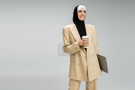 Foto de Mujer de negocios musulmana encantada y de moda con taza de papel y portátil mirando hacia otro lado en gris - Imagen libre de derechos